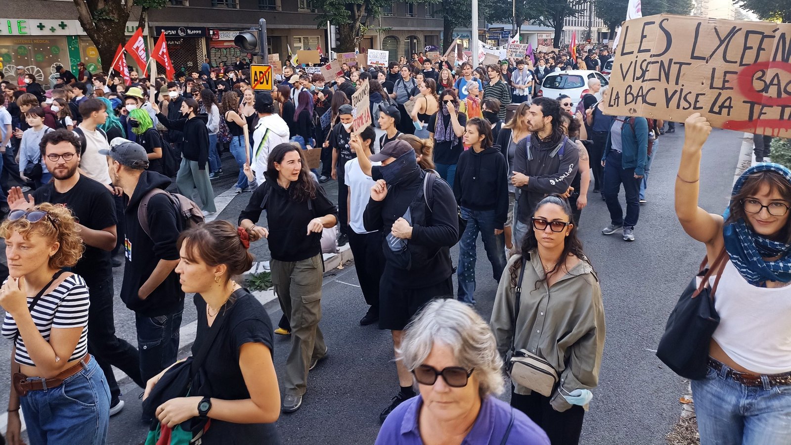 Une foule dense de lycéens et de manifestants défile dans une rue, certains le visage masqué, des drapeaux syndicaux rouges brandis ; au premier plan à droite, une pancarte en carton proclame « Les lycéens visent le bac, la BAC vise la tête ».