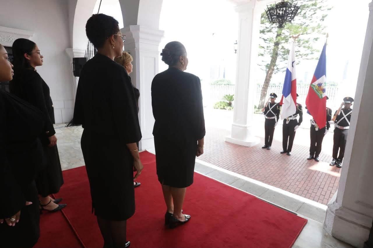 La délégation haïtienne, en tenue sombre, attend sur le tapis rouge à l'entrée du Palais présidentiel panaméen, face aux drapeaux d'Haïti et du Panama et à la garde d'honneur.