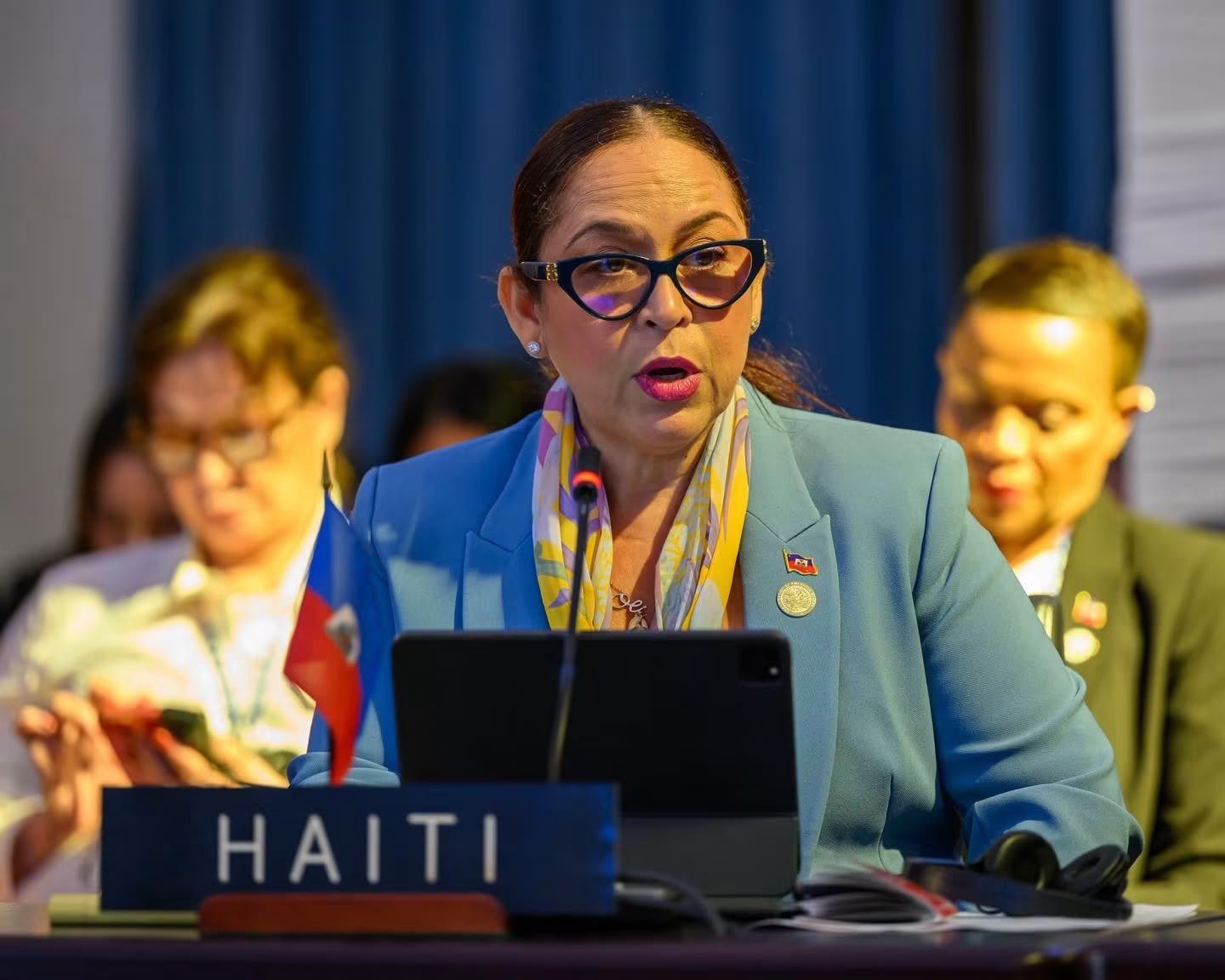 La ministre haïtienne des Affaires étrangères et des Cultes, Raina Forbin, lunettes et foulard coloré, s'exprime au micro derrière une pancarte marquée « HAITI », drapeau haïtien à ses côtés, rideau bleu en arrière-plan.