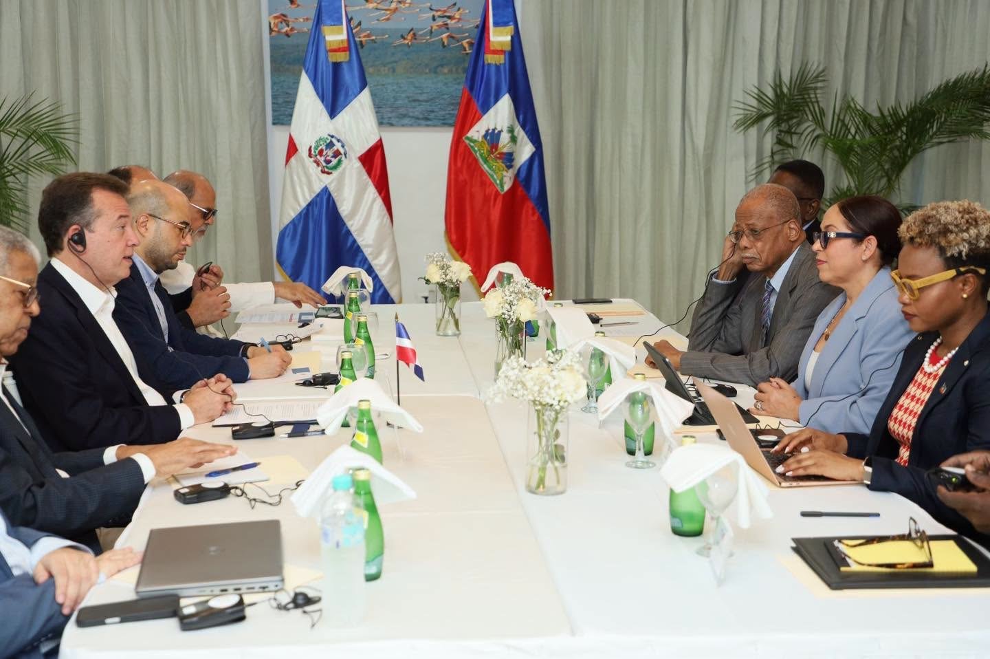 Délégations haïtienne et dominicaine réunies autour d'une table lors d'une rencontre bilatérale à Dajabón, drapeaux des deux pays en arrière-plan.