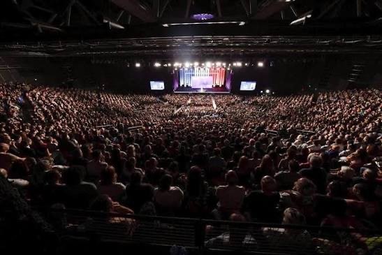 Une foule compacte remplit une grande salle, sous un éclairage de scène rouge et bleu, lors d'un meeting politique.