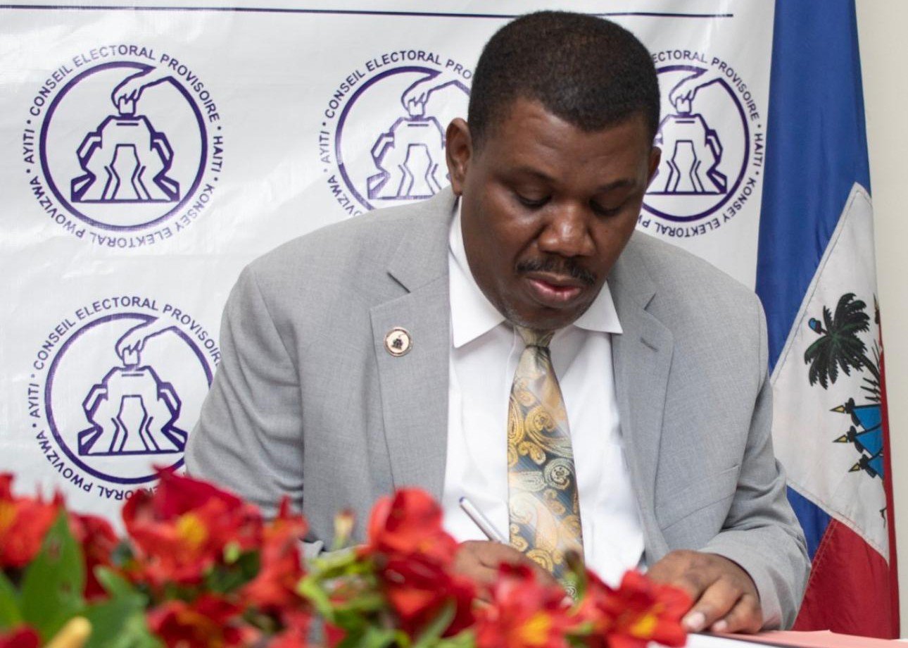Uder Antoine, directeur du Conseil électoral provisoire, signe un document officiel devant la bannière du CEP et le drapeau haïtien.